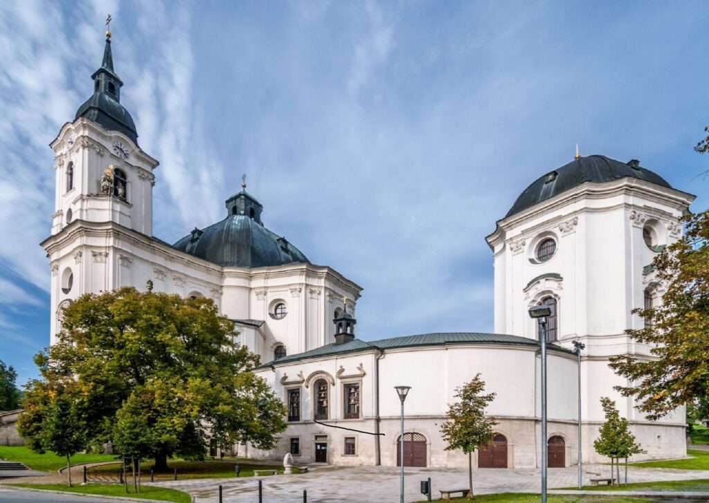 White church with a tall clock tower and copper-green dome, ornate façade, in a tree-lined square against a blue sky.