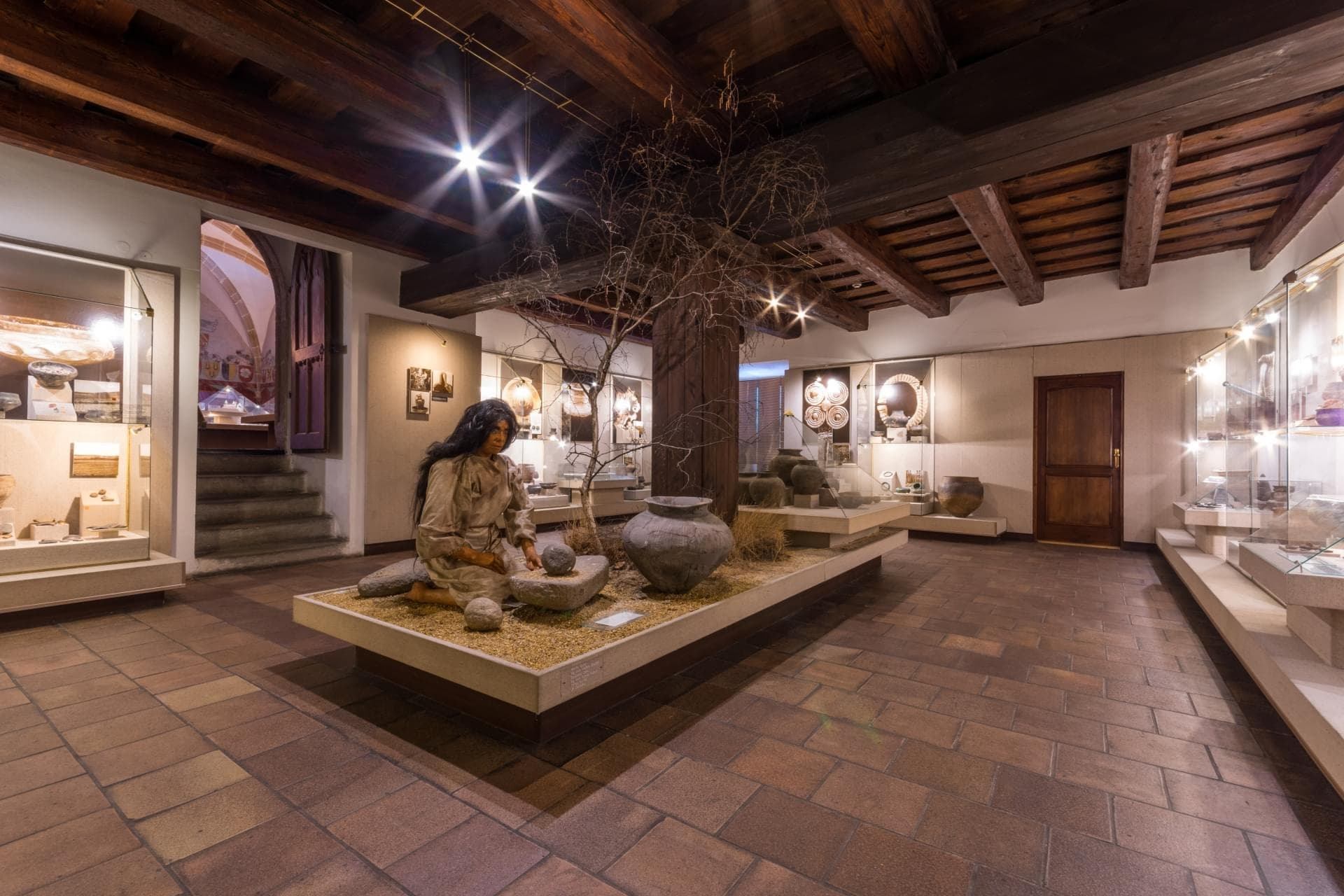 Museum gallery with a life-size seated figure sculpture in a sandy planter, a bare tree, and pottery displays under a wooden-beam ceiling