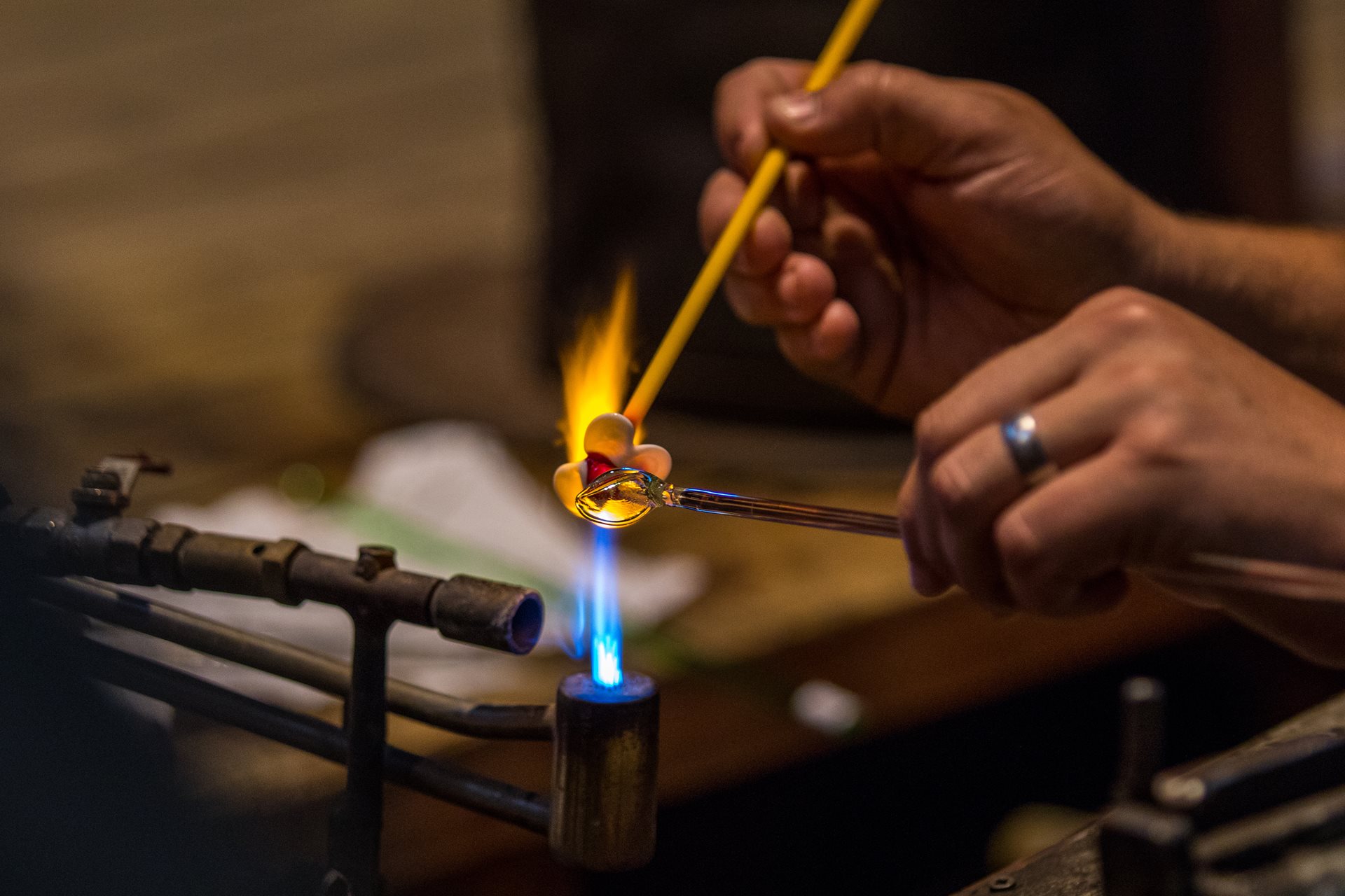Glassblower heats a glowing molten blob on a rod with a torch flame, using a second rod to shape it.