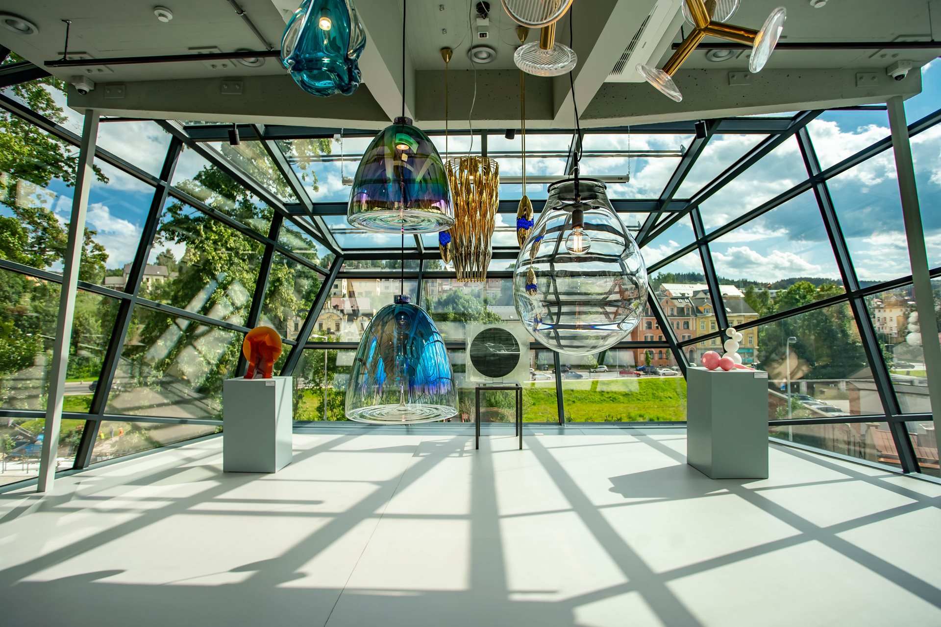 Interior of a bright glass-walled atrium with oversized glass pendant lamps hanging from the ceiling, reflecting light onto the white floor overlooking a cityscape outside