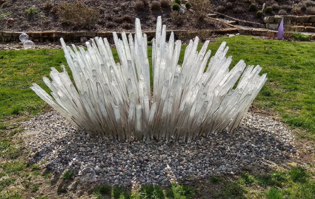 Large cluster of clear glass spikes radiating upward from a gravel bed in a garden. (Outdoor sculpture)