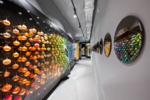 Colorful glass ornaments displayed in a long glass case along a modern gallery hallway, with circular mirrors on the right wall.