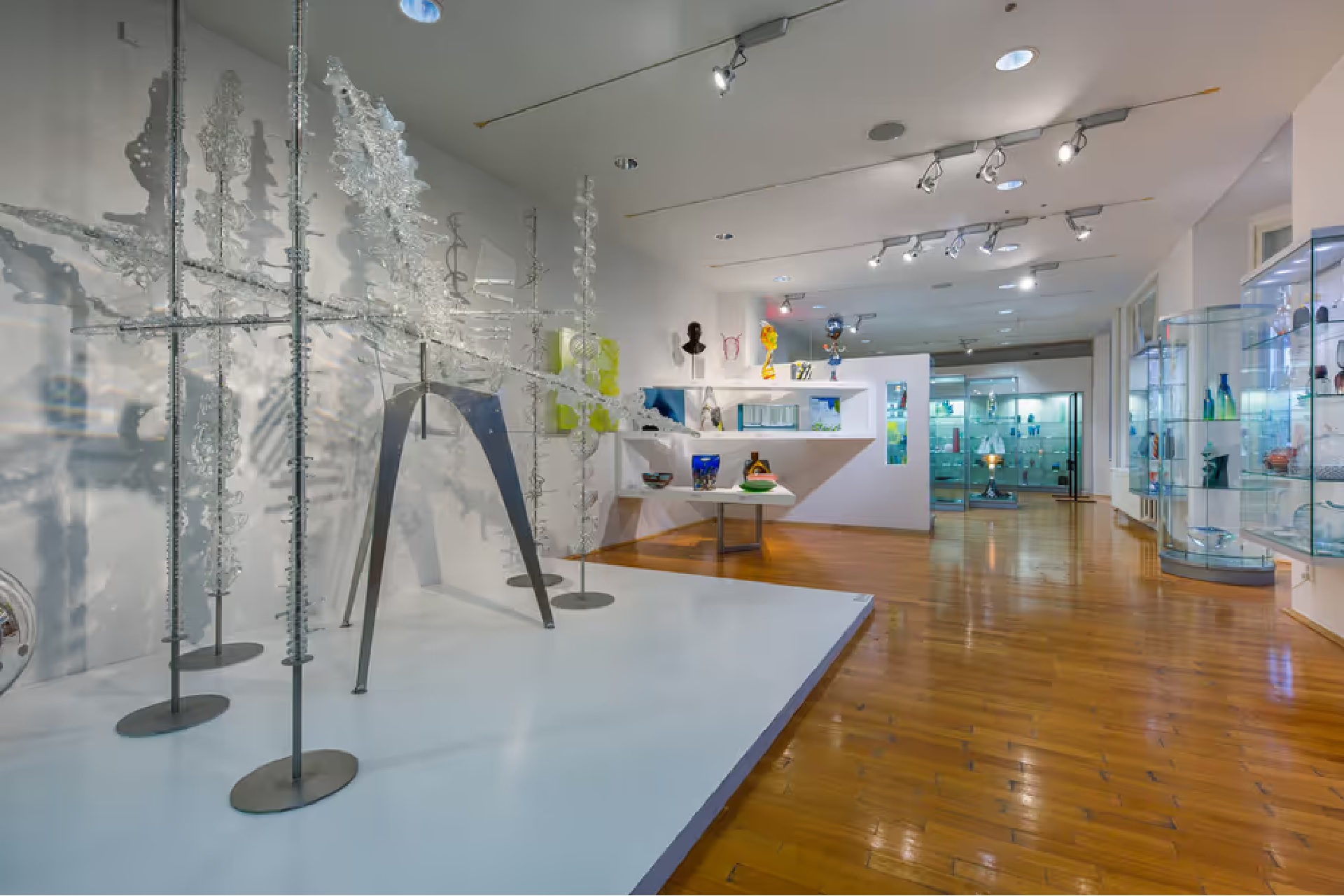 Art gallery interior with tall minimalist sculptures on display along a glass wall and white pedestals, wood floor on the right side.