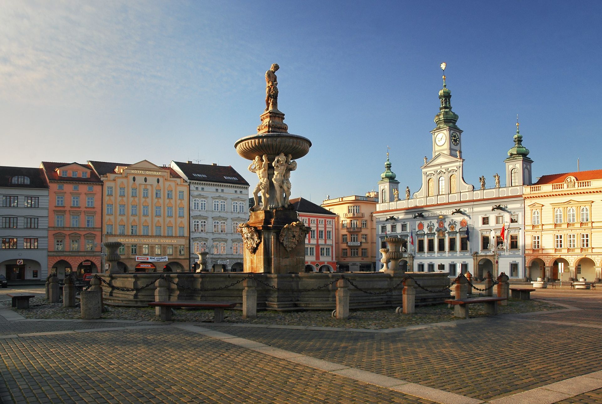 Stone fountain with statue in a sunlit European square, pastel townhouses in the background and a blue sky