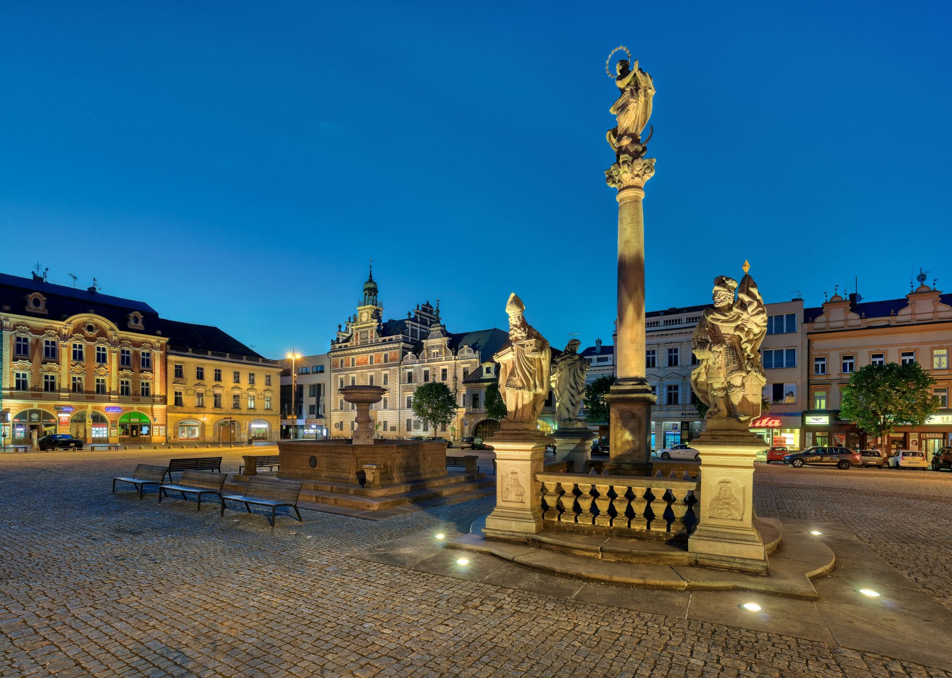 Evening view of a historic square with illuminated statues on pedestals and ornate buildings in the background.