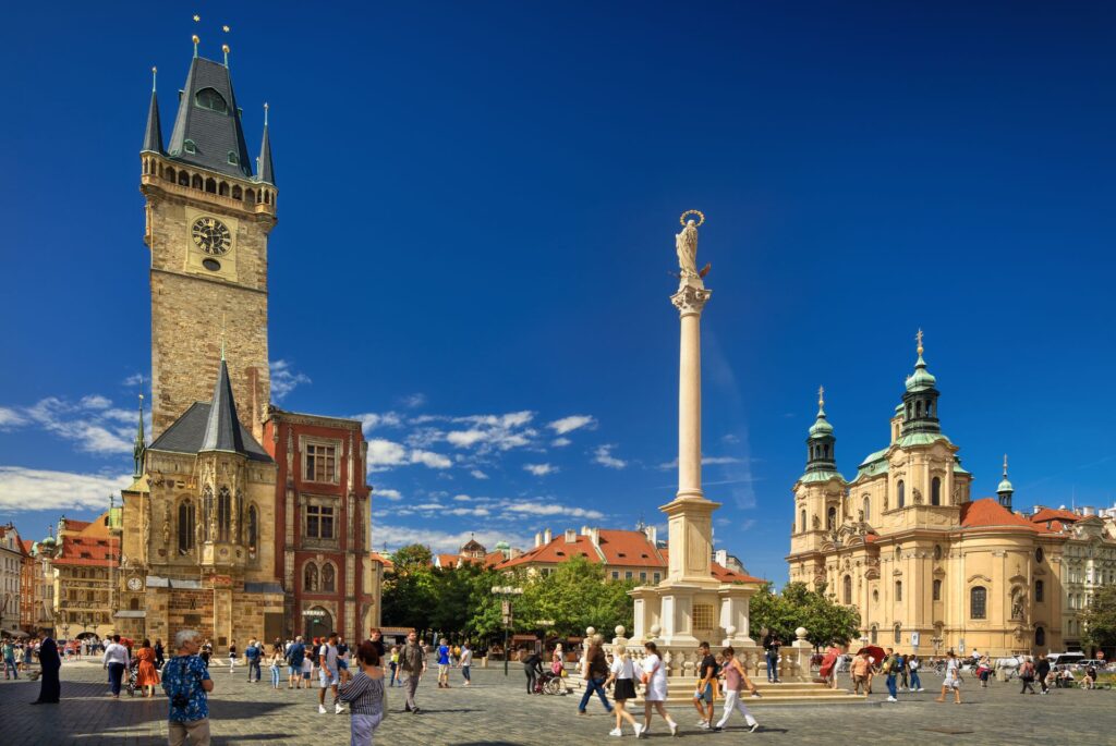 Clock tower in a historic square with a statue on a column and a crowd of people under a clear blue sky.