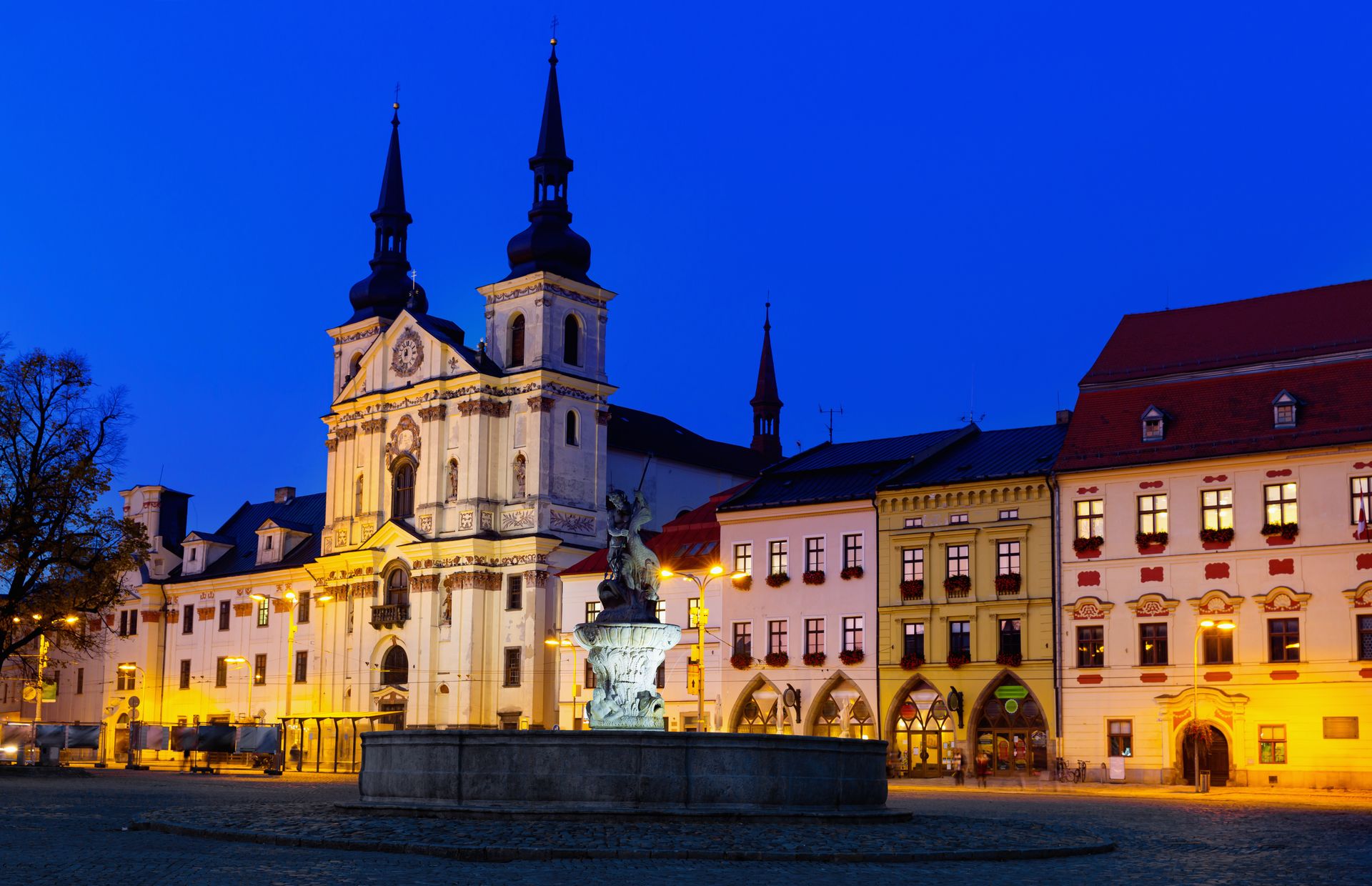 Historic town square at dusk with a grand church façade, twin spires, and a statue fountain in the foreground.