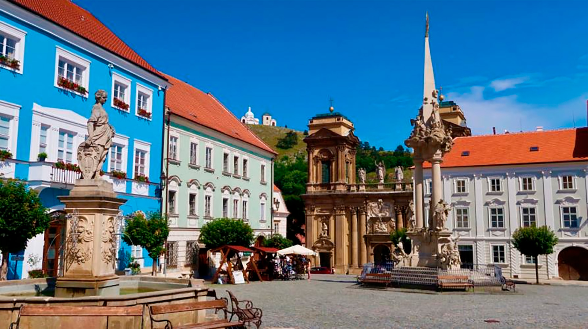 Colorful town square with a bright blue building, ornate fountain with statues, and outdoor cafe under a clear blue sky.