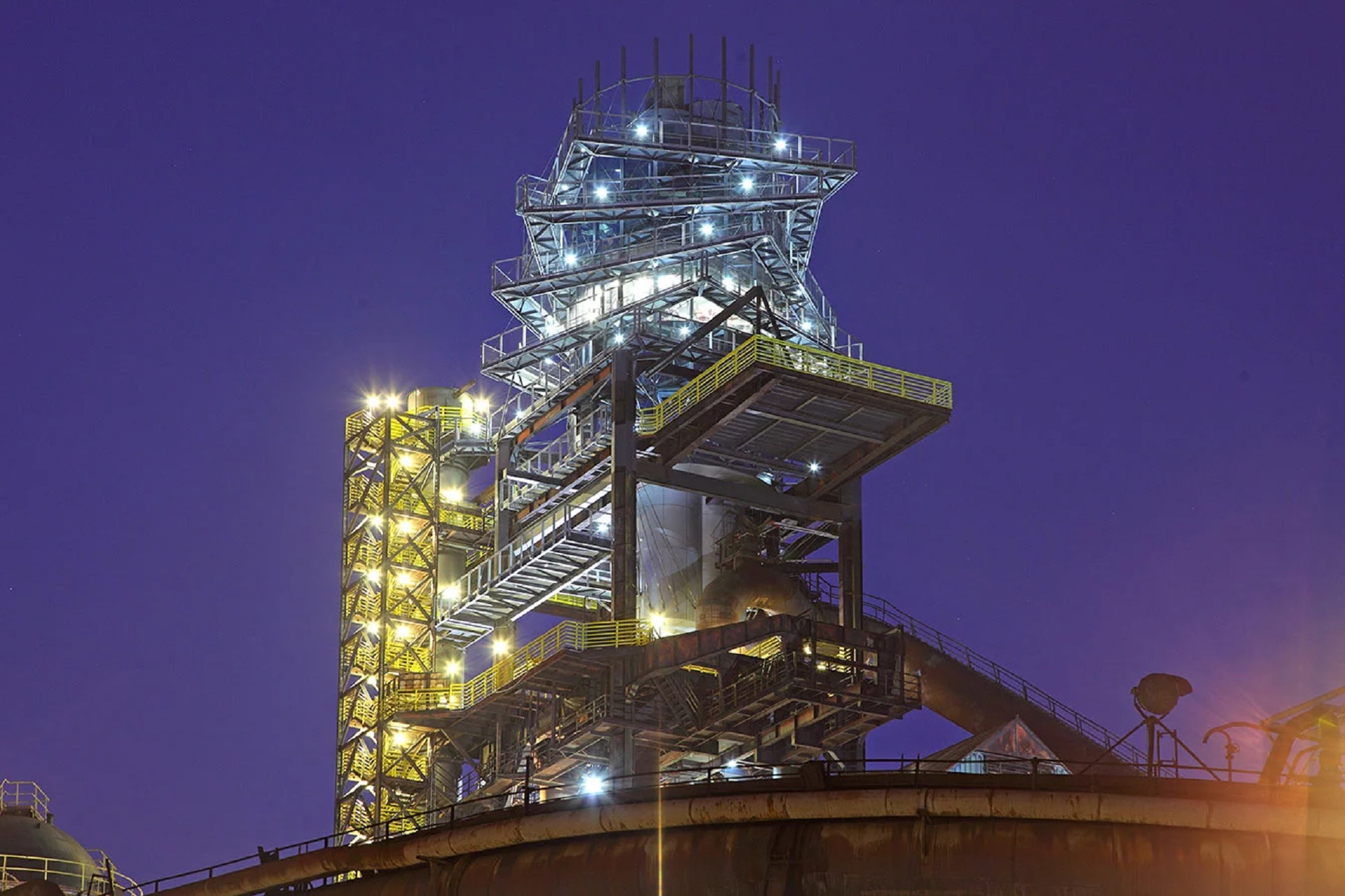 Night view of an industrial refinery tower with illuminated metal walkways against a dark blue sky.