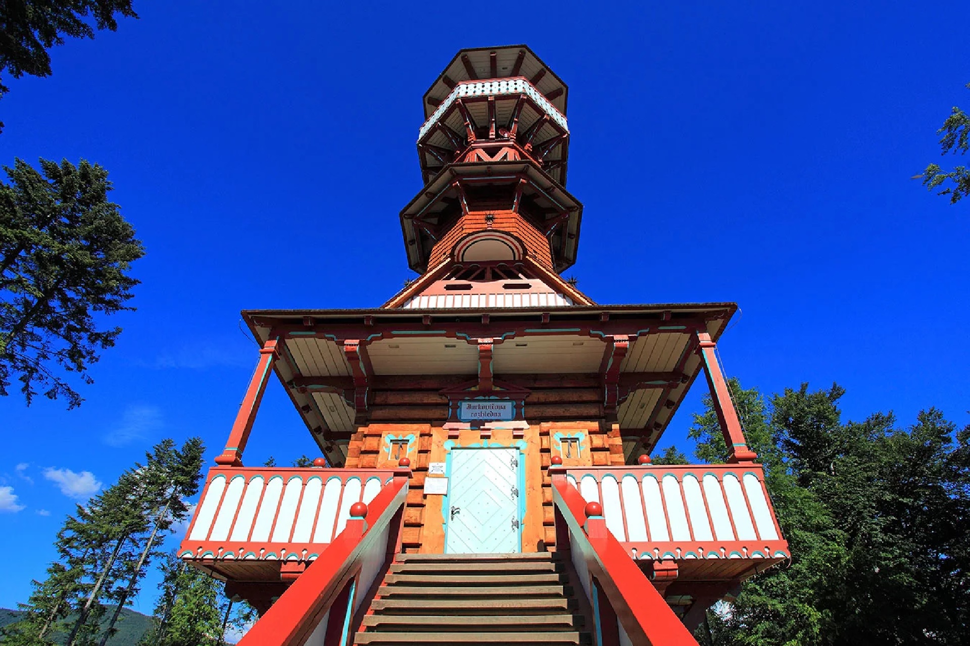 Colorful wooden lookout tower with red railings, reached by a long staircase, set against a clear blue sky and surrounding trees.