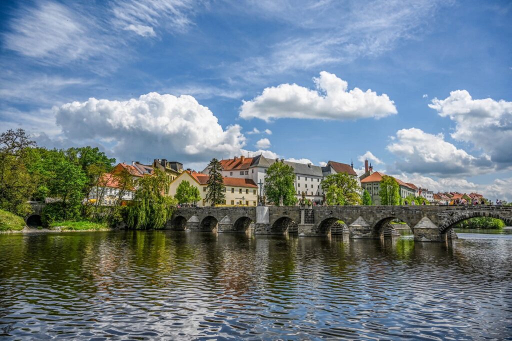 Stone-arch bridge over a calm river with colorful buildings and trees along the bank under a blue sky with clouds, historic town scene.