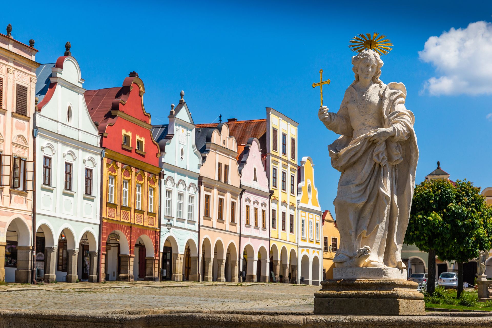 Stone statue with a sunburst halo and cross held aloft in a square, colorful historic buildings behind under a blue sky, archways at street level, sunny day.