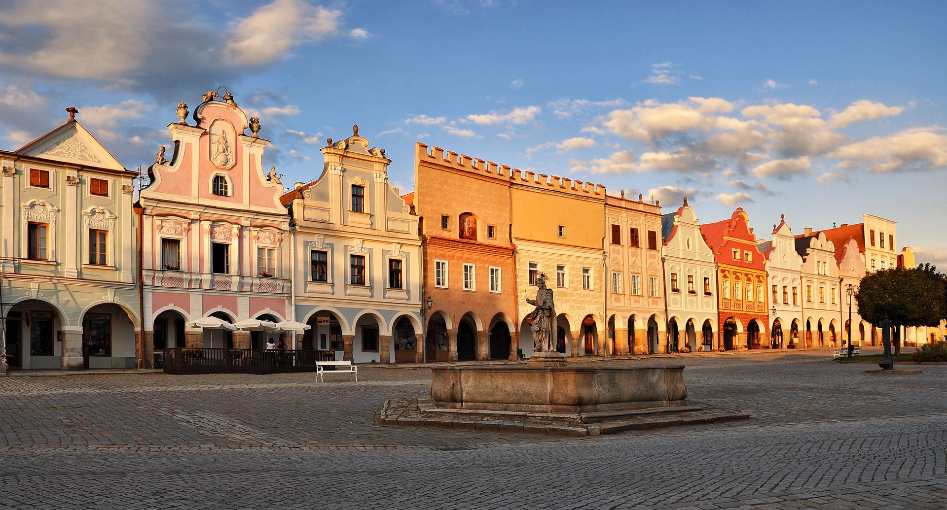 Row of pastel historic buildings with arches along a cobblestone square and a central fountain statue in the foreground.