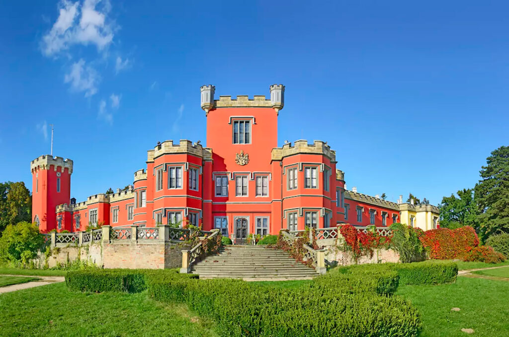 Red castle-like mansion with crenellated towers and symmetrical windows, set against a bright blue sky with manicured grounds and stairs leading to the entrance.