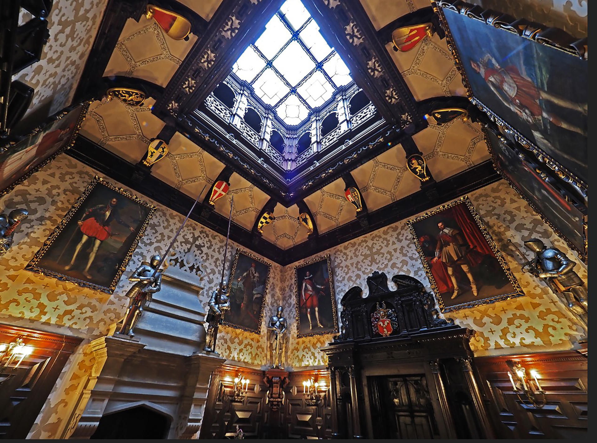 Interior of a medieval-style hall with a glass skylight, heraldic banners, portraits, and suits of armor along dark wood panels.
