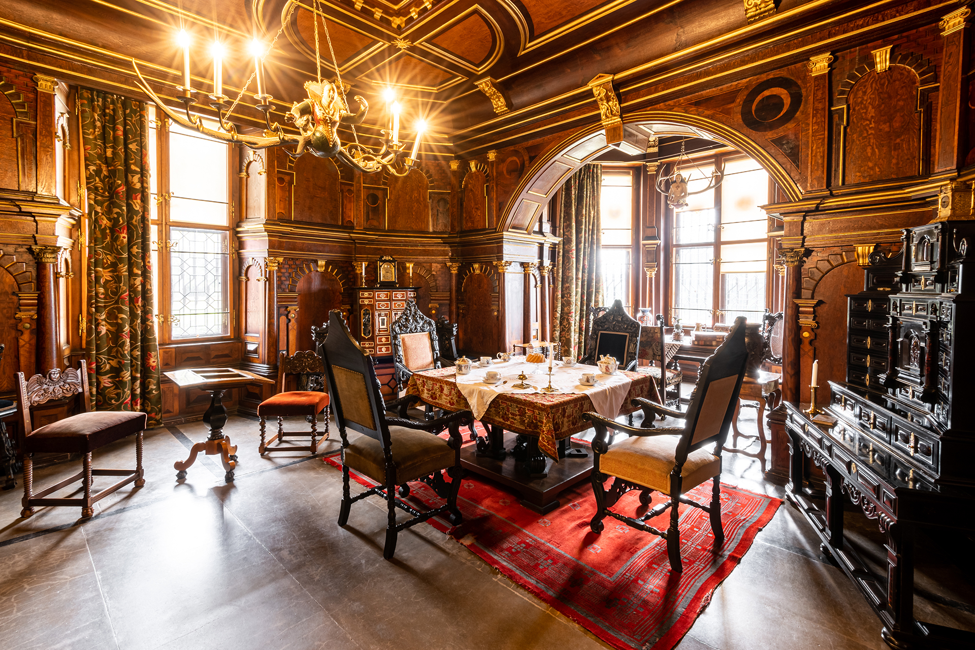 Luxurious wood-paneled dining room with a chandelier, ornate arches, and a table set with china for tea or dinner.