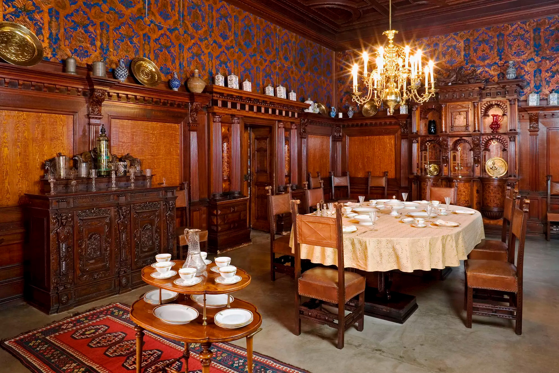 Elegant dining room with dark carved wood paneling, ornate cabinets, and a chandelier above a round table set for tea.