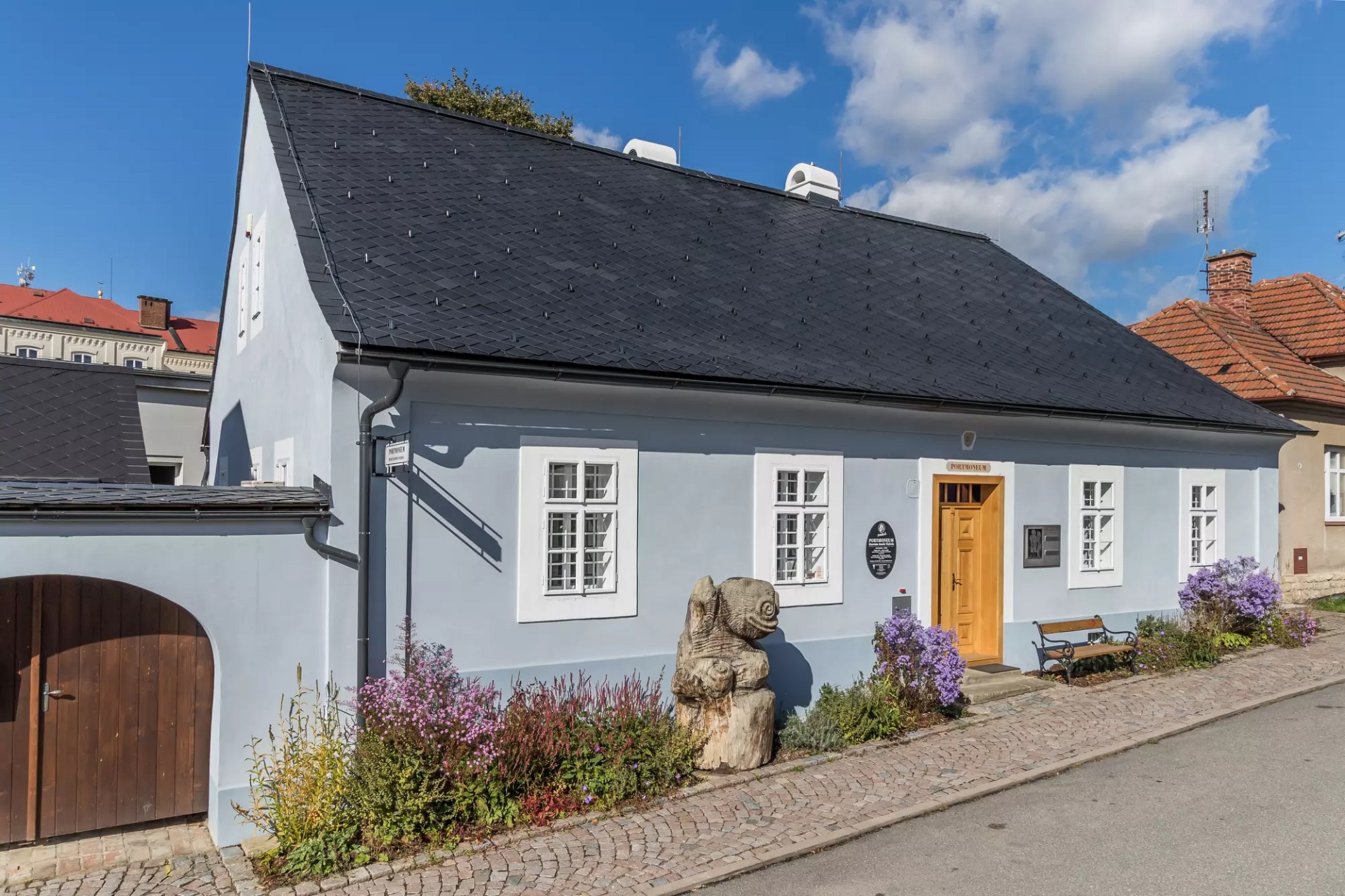 Pale blue, single-story building with a dark tiled roof, white-framed windows, and a wooden door, surrounded by a small garden of pink-purple flowers and a stone sculpture near the sidewalk.