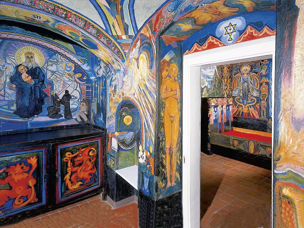 Colorful muraled corridor with religious figures, including a Madonna and child on the left, leading to a doorway into a rooms of vibrant art.