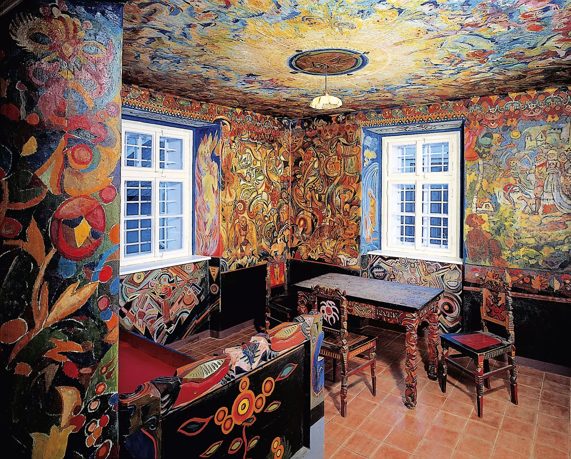 Room interior with bright, swirling mural patterns covering walls and columns, blue window frames, and carved wooden benches and tables under a hanging light.