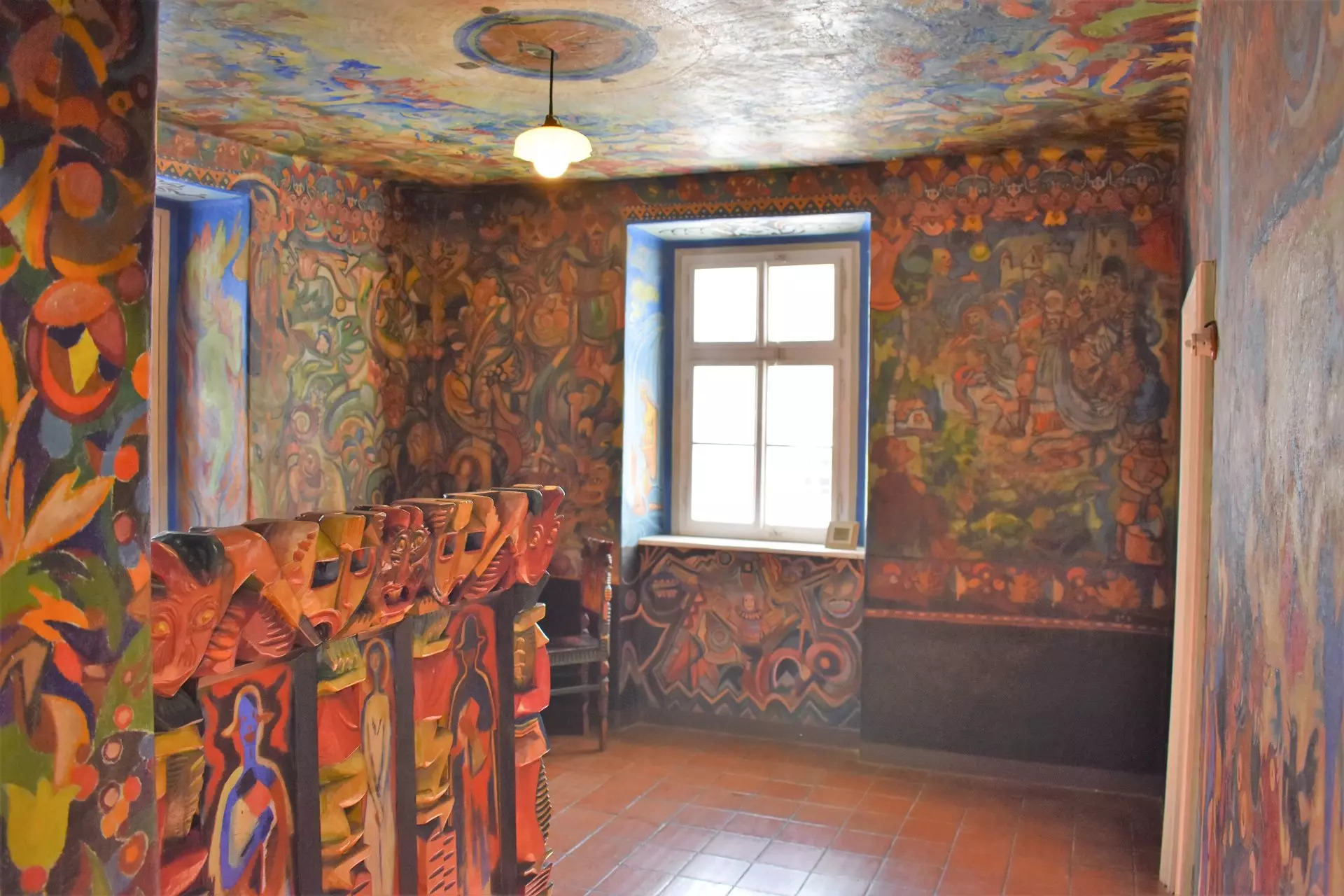 Whimsical room with vibrant mural walls and rows of orange-painted chairs facing an unseen stage, near a window and hanging ceiling light.