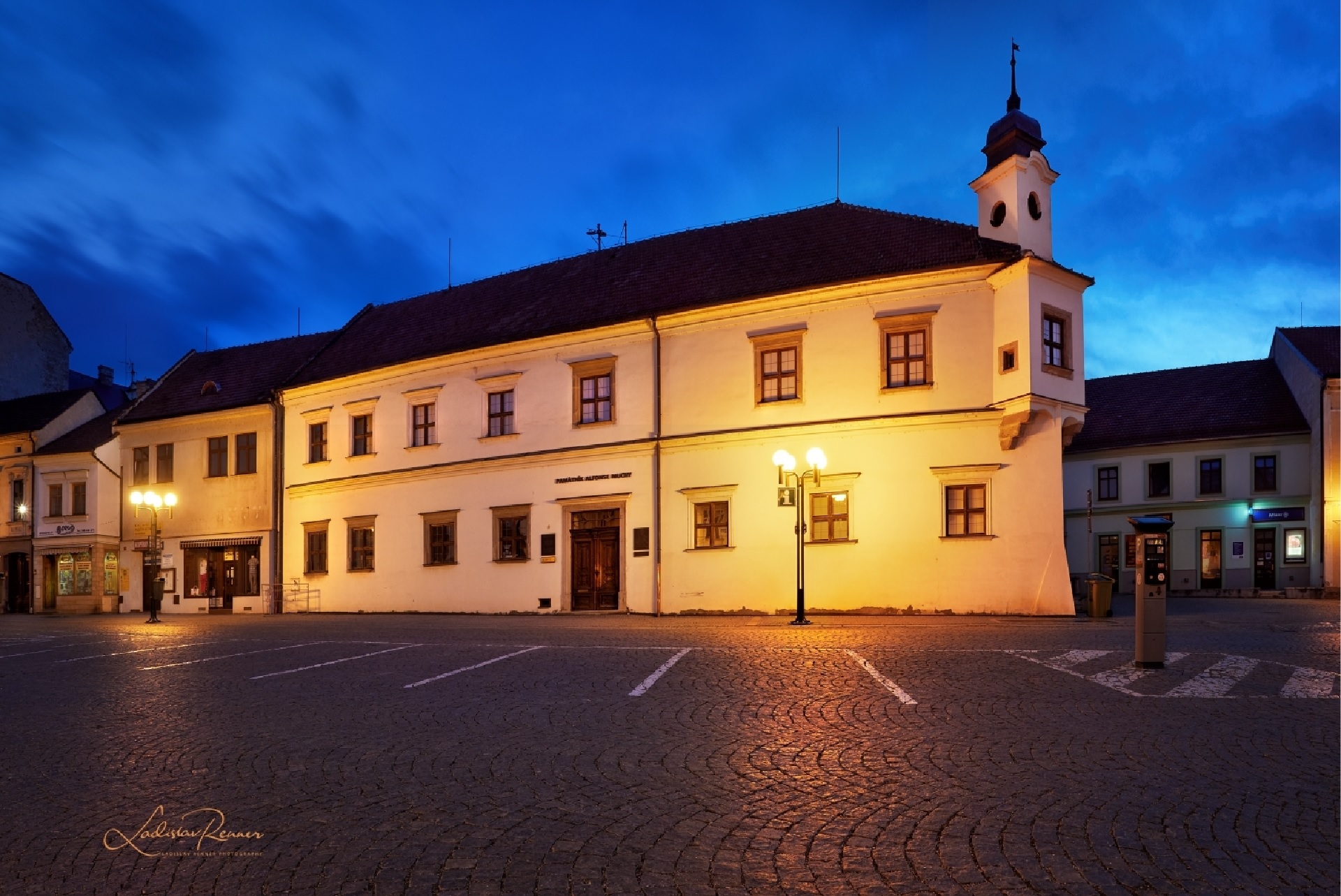 White, two-story historic building with a small tower at the right, illuminated by warm streetlights in a cobblestone town square at dusk, blue sky above.