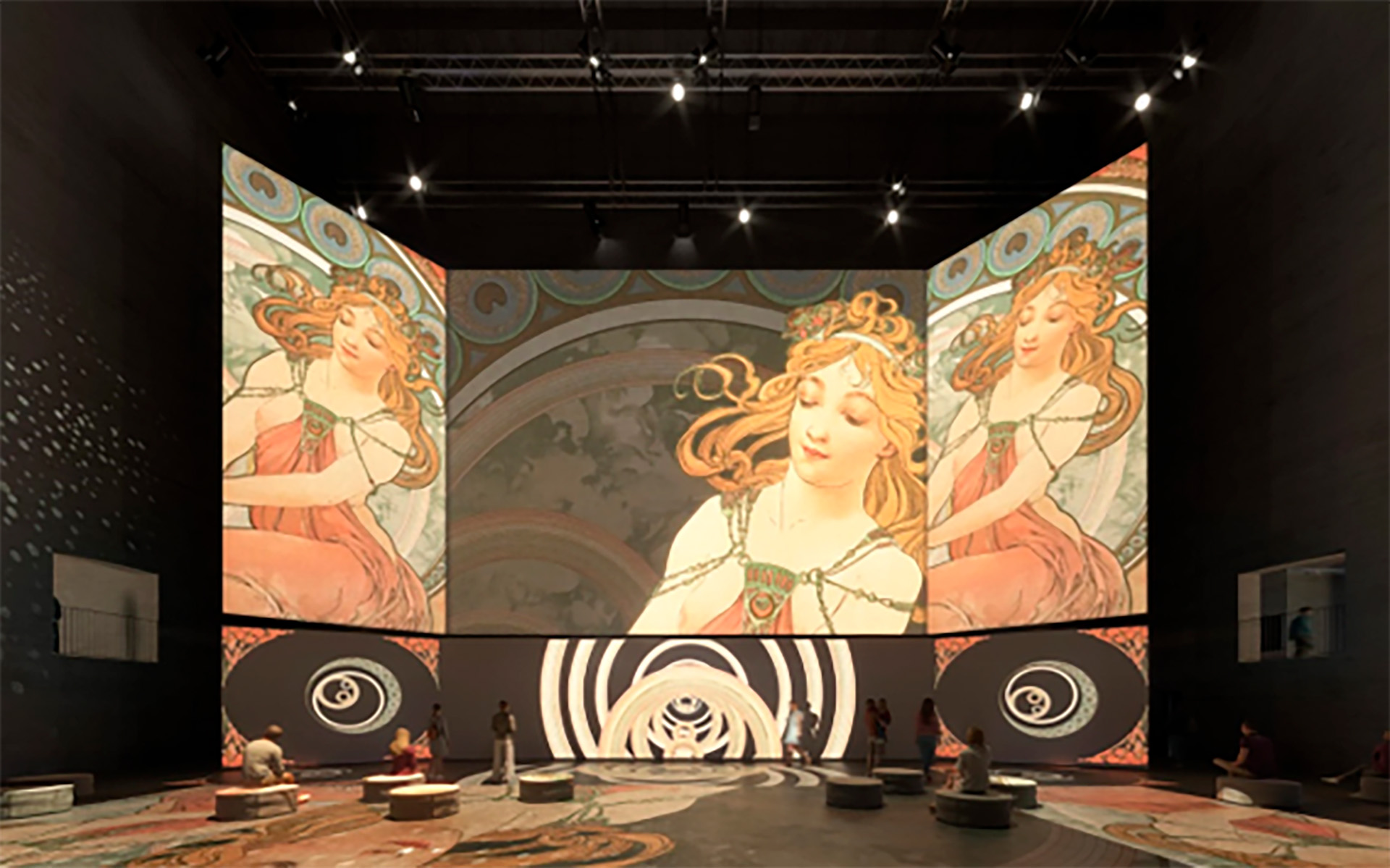 Indoor gallery or exhibit space featuring a large multi-panel mural of three angelic women with flowing hair, surrounded by circular decorative patterns and rings on the floor, with visitors seated on small circular platforms.