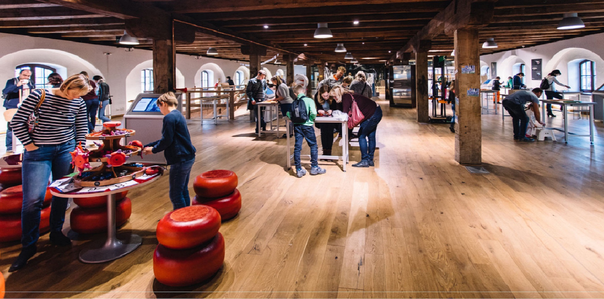 People of various ages engaging with hands-on exhibits in a spacious museum-like gallery with wooden floors and exposed beams.