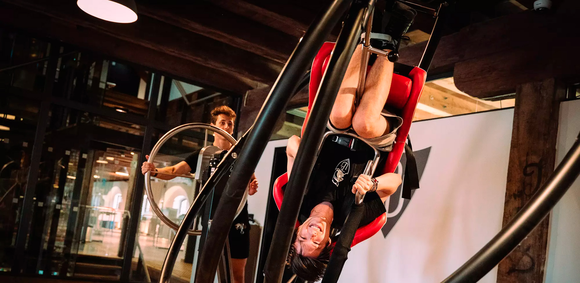 Person suspended upside down in a red padded harness on a large circular fitness apparatus, with a trainer nearby in a gym.