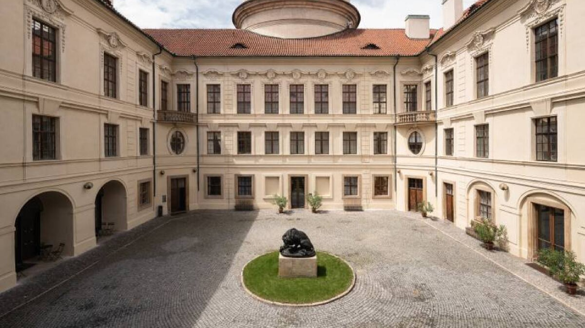 Historic courtyard with a circular grassy island and a fountain statue in the center, surrounded by pale buildings with arched doorways and numerous windows.