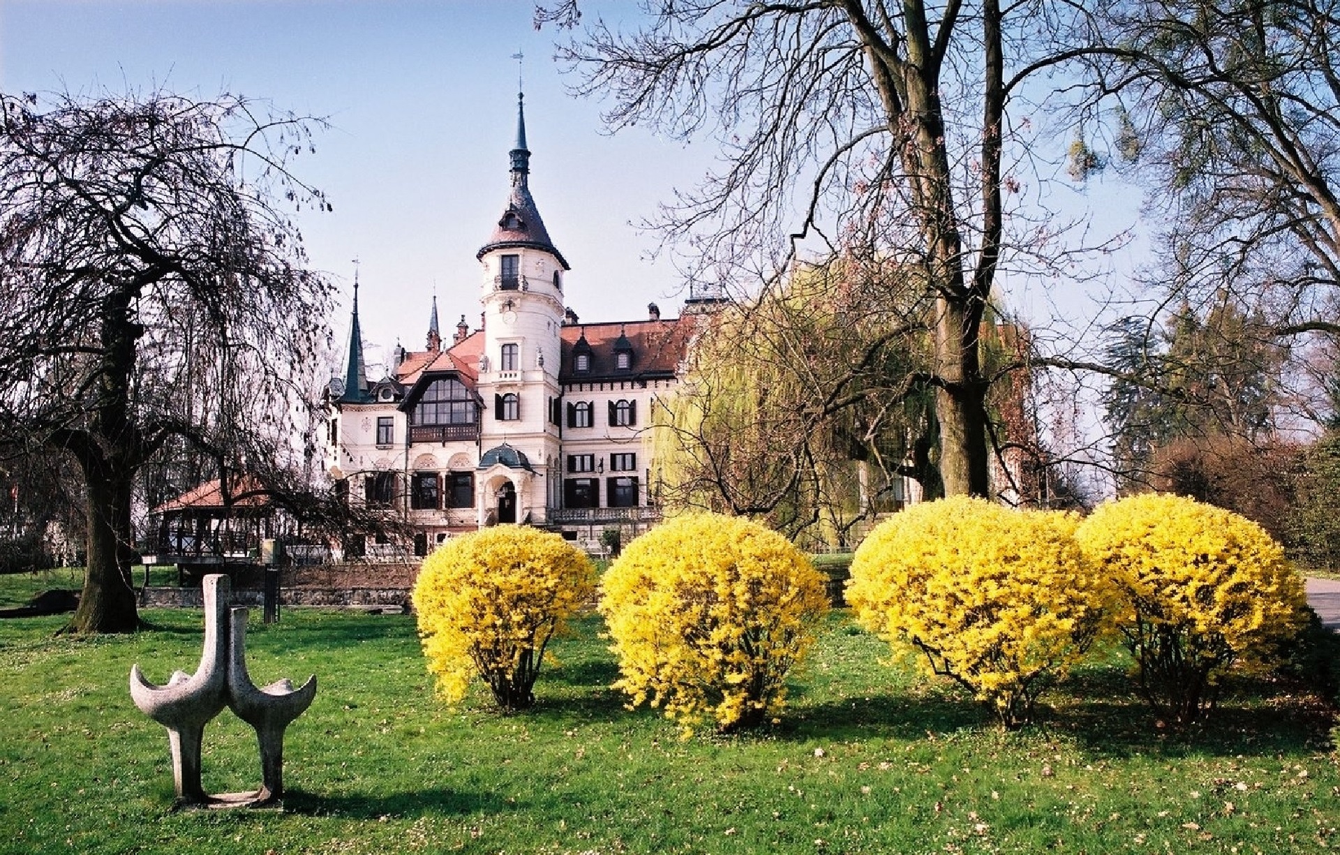 Castle-like building with tall tower, framed by bare trees, in a landscaped garden with bright yellow bushes in the foreground.