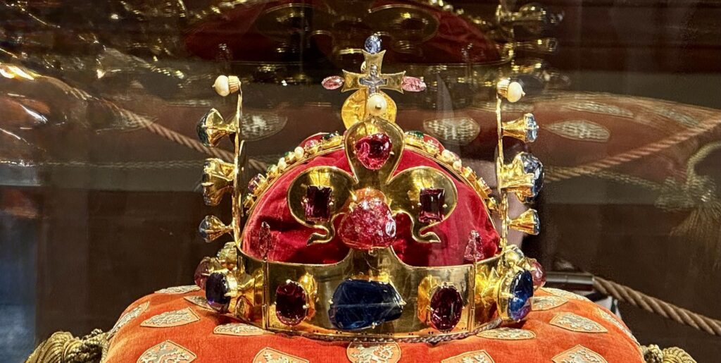 Golden crown encrusted with red and blue gems on a red velvet cushion inside a glass display case.