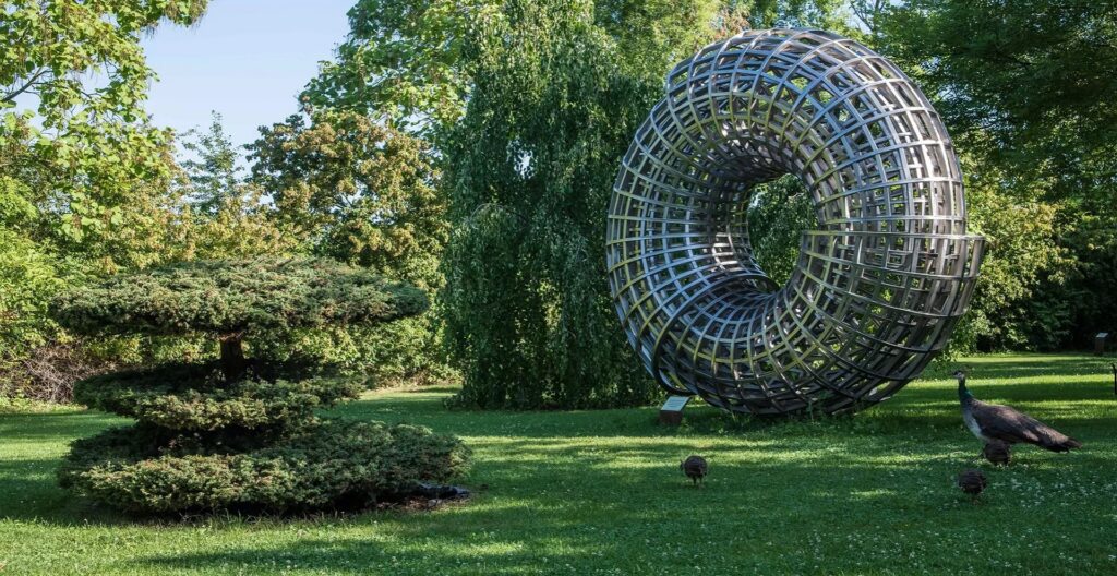 Large metallic circular lattice sculpture in a sunlit park with green grass and trees, with a peacock nearby for scale and life.