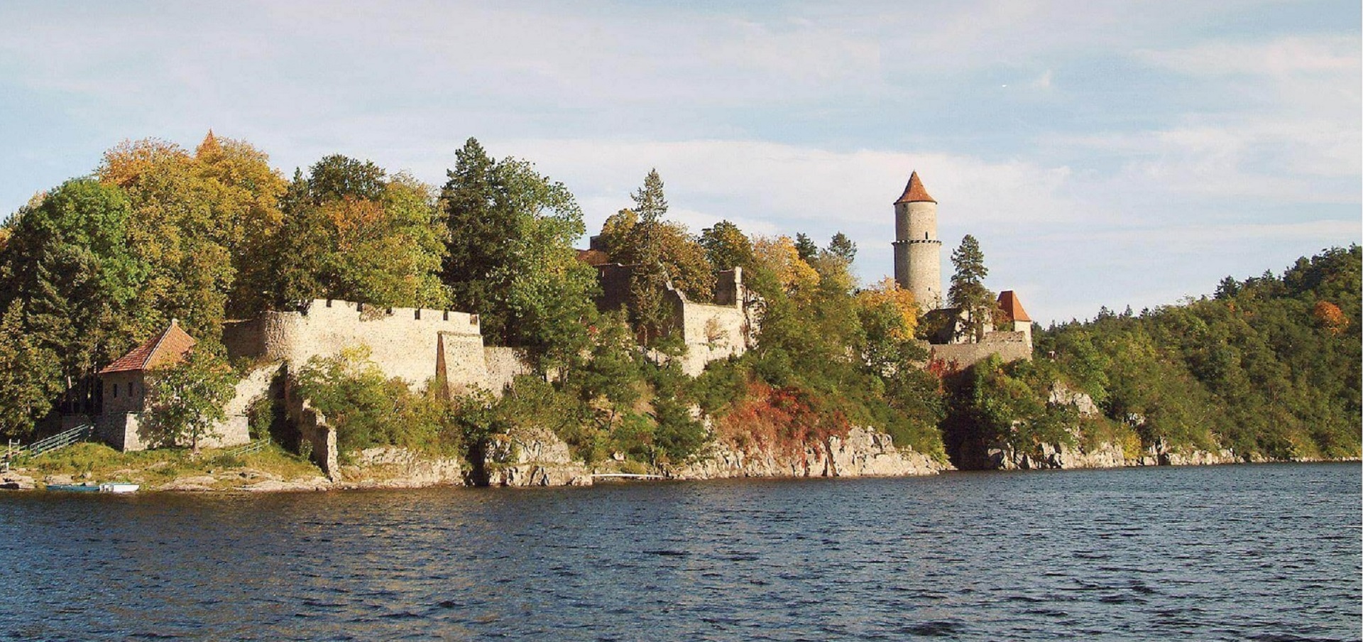 Stone fortress on a rocky shoreline, with a tall round-topped tower and autumn trees forming a backdrop by a calm lake; blue sky above.