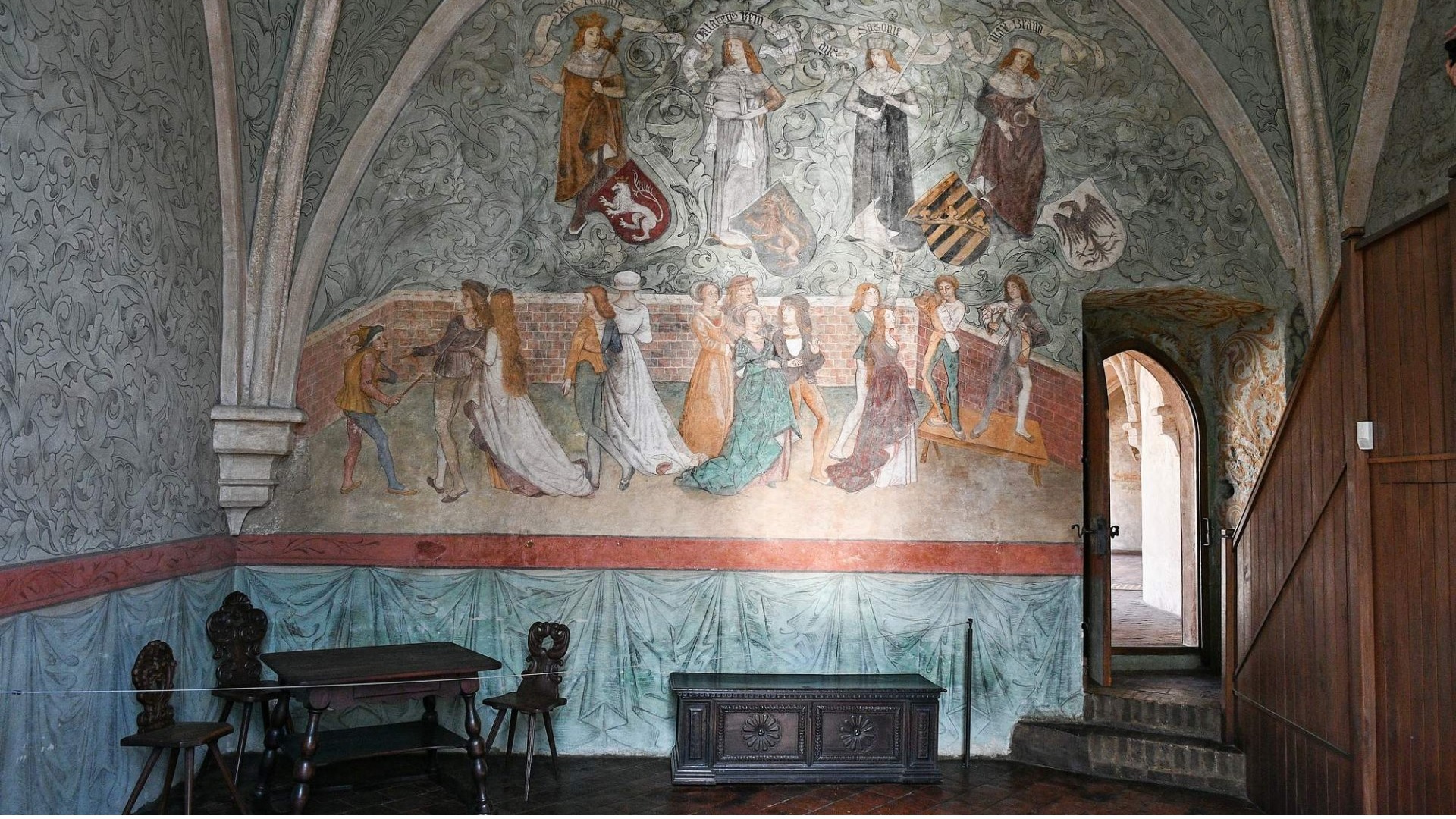 Historic room with a large wall mural depicting nobles and villagers and coat-of-arms under arched, vine-patterned ceiling.