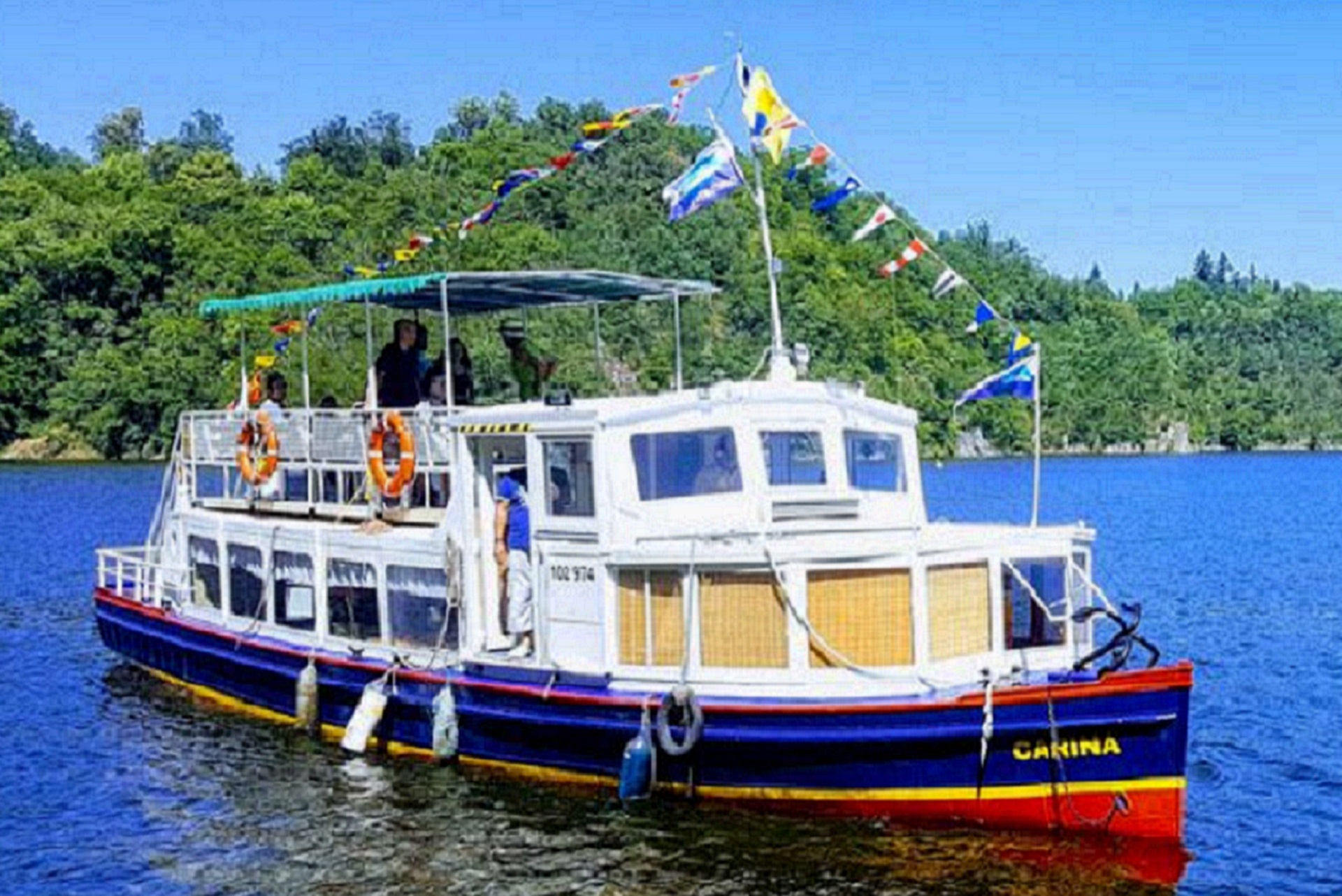 Passenger boat named Carina on a blue lake, with people on deck and colorful flags fluttering above; forested shoreline in the background.