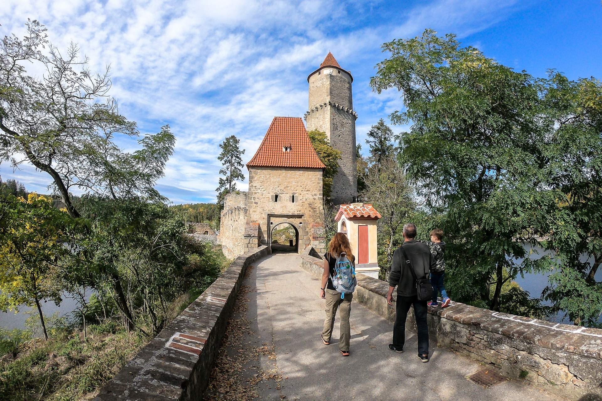 Stone fortress with a tall round tower and red-tiled roof at a gateway, as visitors walk along a stone path lined with trees over a river.