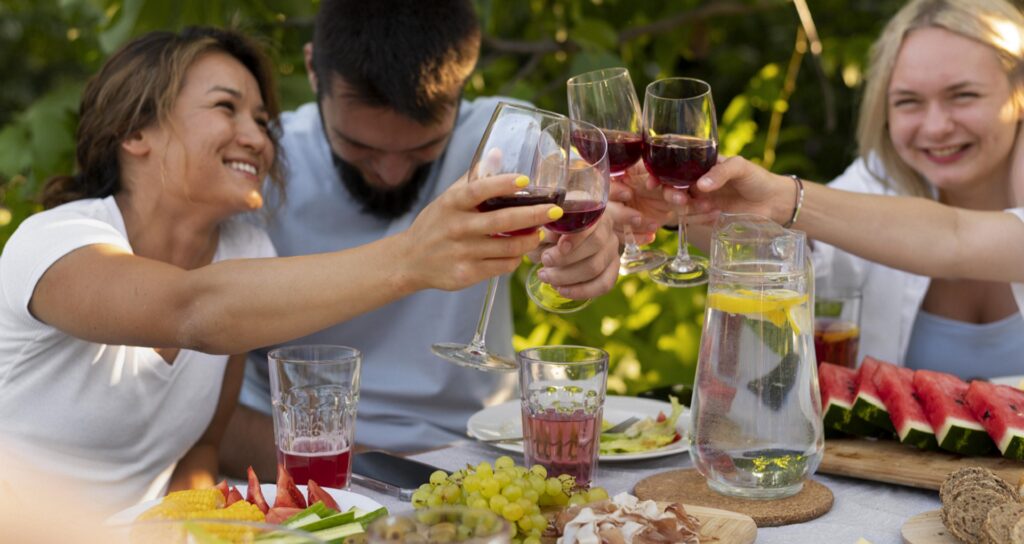People raising glasses in a cheerful outdoor toast around a table with watermelon and fruit platters.