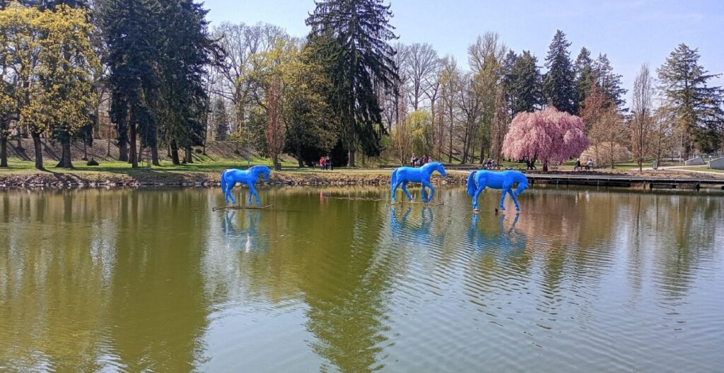 Three blue horse statues standing on submerged platforms in a park pond, with green trees and a pink blossoming tree in the background.