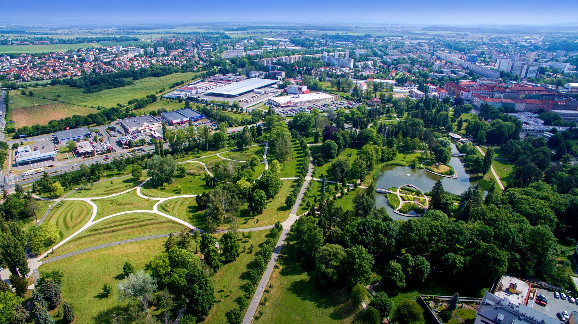 Aerial view of a city park with winding paths, a central pond, and surrounding urban buildings and greenery.