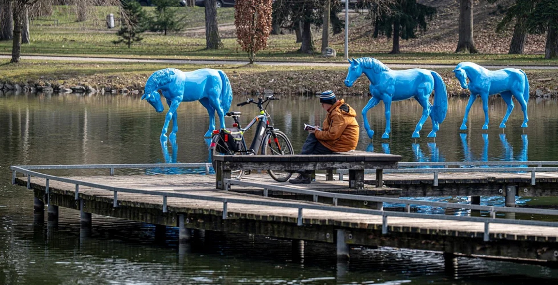Man in an orange jacket sits on a dock by a pond reading a book, with bright blue horse sculptures in the water and a bicycle nearby.