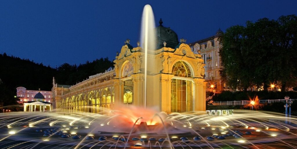 Vista noturna de um palacete estreito, com uma praça repletas de pequenos chafarizes iluminados que esguicham fios de água, formando efeito de leito de rio.