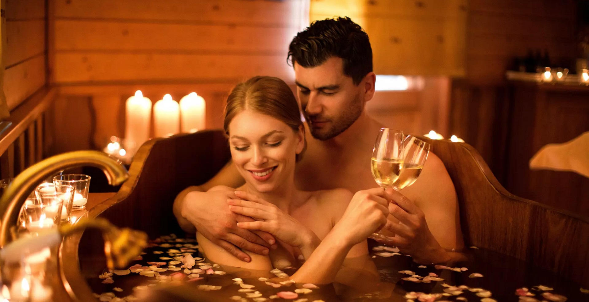 Casal sorridente em uma banheira de madeira, visivelmente em ambiente de spas. Na água da banheira flutuam ervas e pétalas de flores. Ao lado esquerdo e atrás da banheira há aparadores e sobre eles, velas acesas.