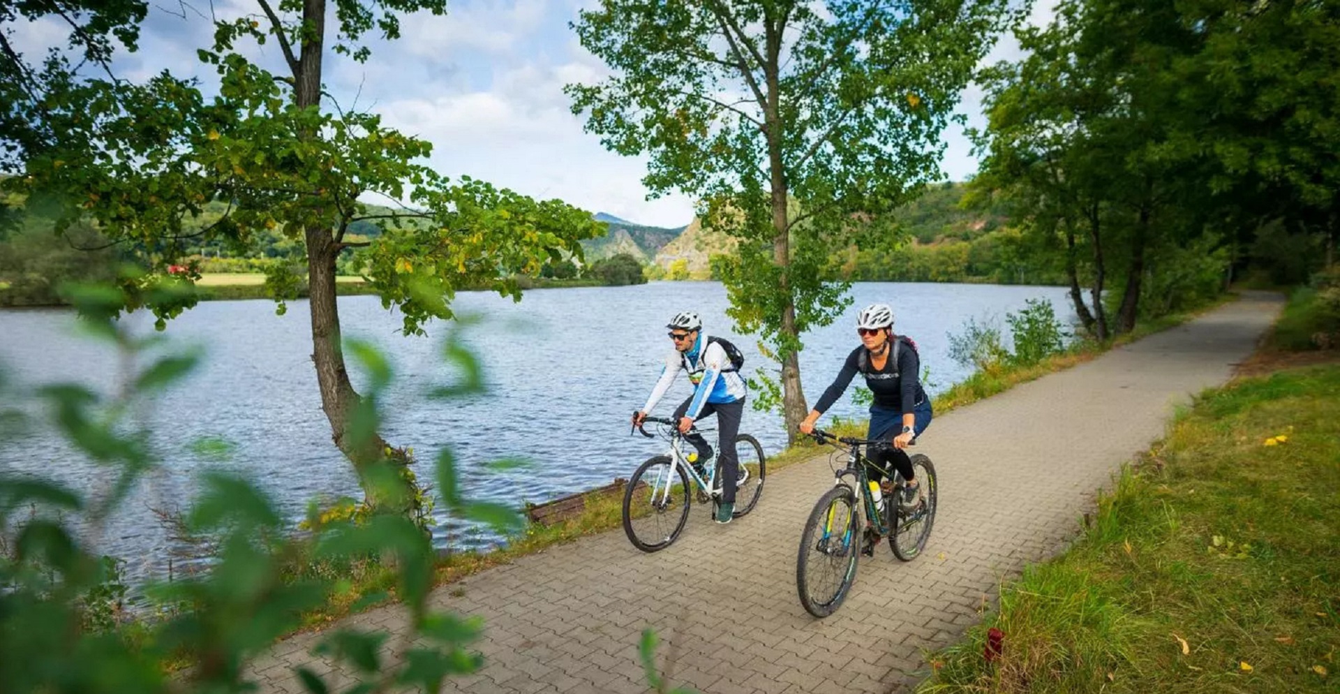 Casal pedala em trilha ciclística na margem de um rio. O trecho tem belas árvores.