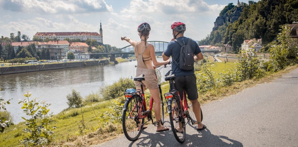 Casal segura bicicletas em uma trilha de ciclismo na margem de um rio. A mulher aponta para a margem oposta, onde há uma igreja e outros prédios.