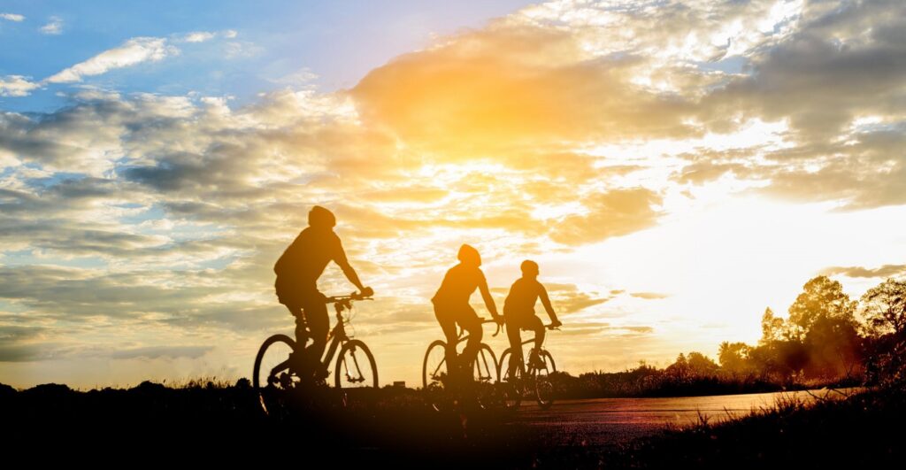 Um amanhecer com raios de Sol dourando as nuvens e silhuetas de três ciclistas.
