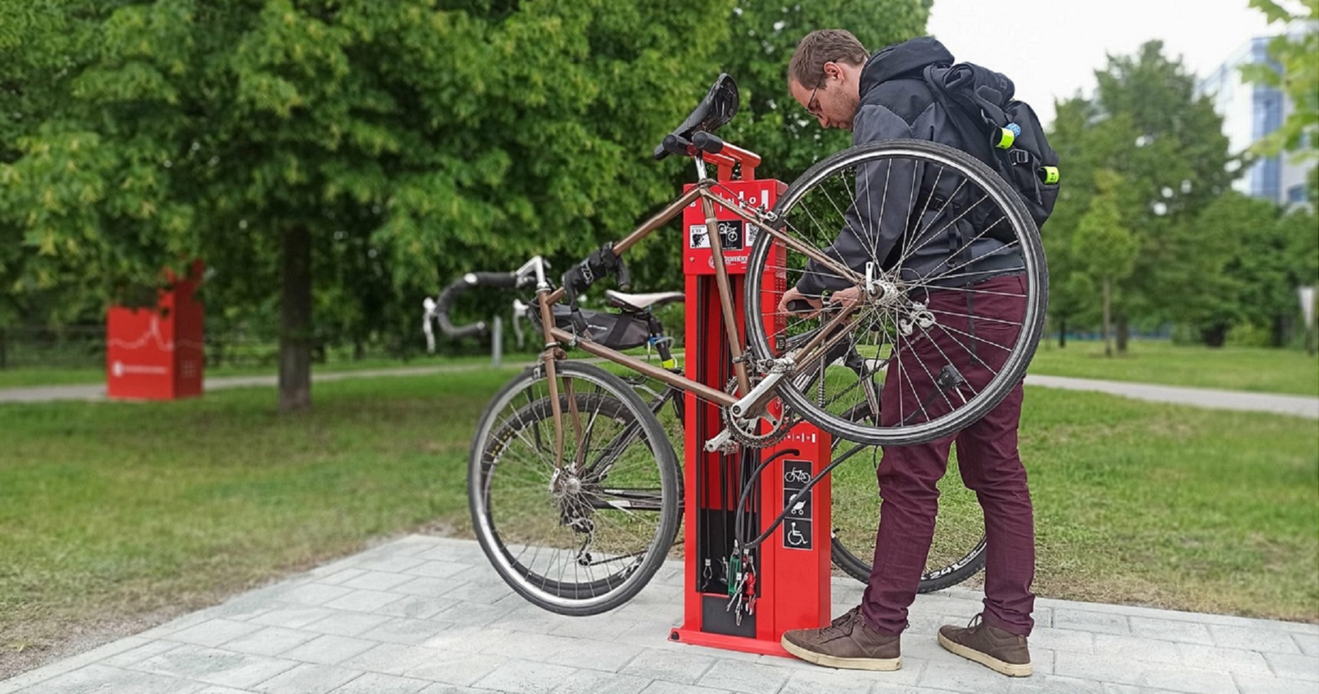 Em primeiro plano, um ciclista com capacete esta´junto a um totem vermelho, utilizando ferramenta presa no interior do totem para reparar a bicicleta