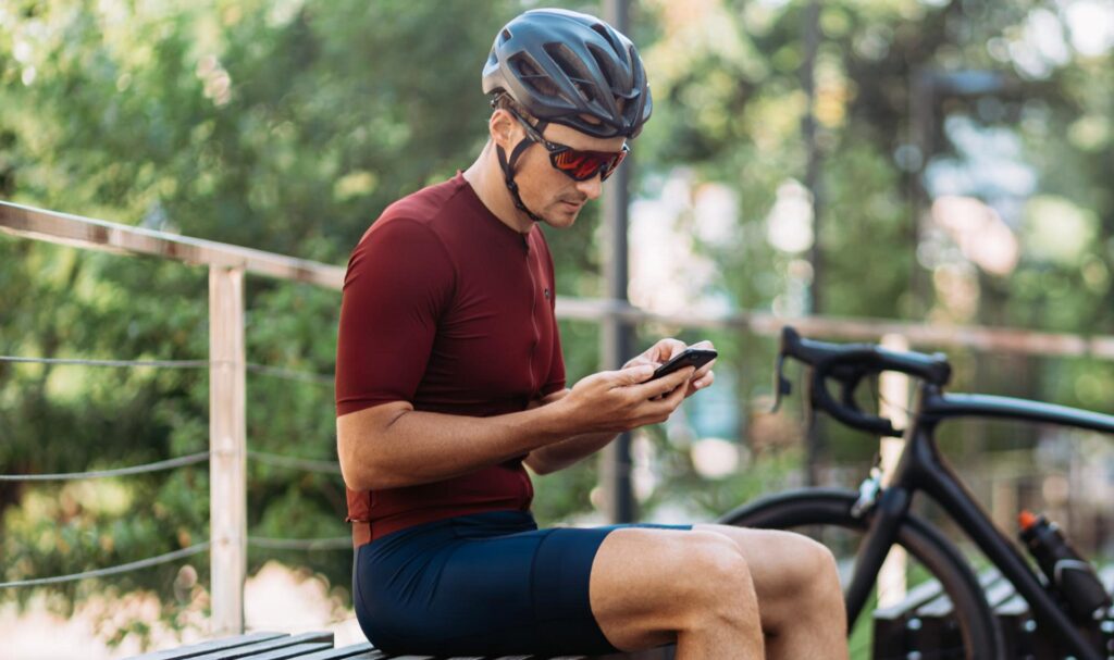 Homem com capacete de ciclista e junto a uma bicicleta olha para a tela do celular.